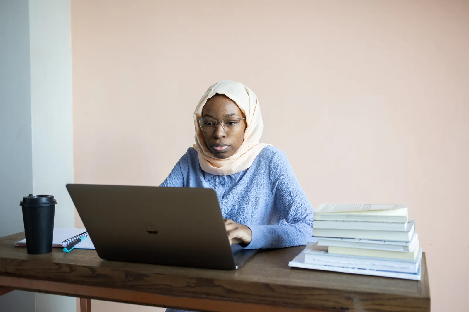 Voksen kvinne med hijab og briller arbeider på bærbar PC ved en bokstabel